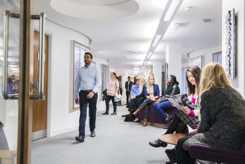 View looking down a hallway with doors on left and students sitting along benches on left